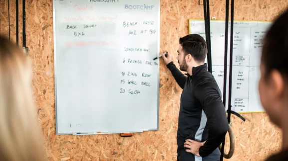 Προπονητής γράφει το πρόγραμμα της ημέρας (WOD) σε λευκό πίνακα γυμναστηρίου – Οι 5+2 άγραφοι κανόνες του γυμναστηρίου για να μην είσαι «αυτός» ο τύπος από το Gymscouter.